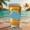 Glass of beer with "Lake Monroe" and a blue shape, on a beach at sunset.