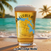 A tall glass of beer with foam sits on a sandy beach.