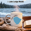 White mug with "Lake Mary Minnesota" design sits by rainy window, knit blanket and book.