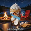 Glass of iced coffee with whipped cream and s'mores ingredients by a campfire.
