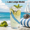 Glass with drink, limes, mint, and compass design on a beach with a sailboat.