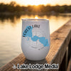 A white insulated wine tumbler featuring a blue lake map and compass graphic sits on a wooden dock at sunset.