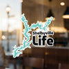 Turquoise Shelbyville life decal with text on a dirty window with blurred background.