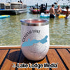 A white stemless tumbler with a Geneva Lake Wisconsin map design rests on a wooden boat deck with people swimming in the background.