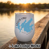 A white stemless wine tumbler rests on a wooden dock with a lake sunset background.
