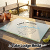 A rectangular glass cutting board rests on a wooden dock, featuring a Lake Hickory compass design.
