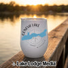 A white stemless wine tumbler with a blue map of Geneva Lake sits on a wooden dock at sunset.