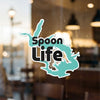 "Spoon Life" sticker on a window with blurred background.