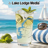 Glass with drink, limes, and mint on beach with sailboat and sunglasses.