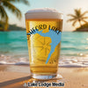 Glass of beer with "SHFORD LAKE" and a compass design on a beach.