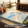 A glass cutting board featuring a map of Saratoga Lake and a compass design sits on an outdoor wooden table near a dock.
