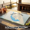 A glass cutting board featuring a blue map of Sanibel Island, Florida, and a compass rose graphic, resting on a wooden dock.