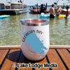 A white insulated wine tumbler with a Georgian Bay Ontario design rests on a wooden boat deck overlooking a sunny lake with boats and swimmers.