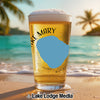 Glass of beer with "Lake Mary" and compass graphic on a beach at sunset.
