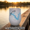 A white stemless wine tumbler rests on a wooden dock beside a lake at sunset.