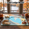 A glass cutting board featuring a blue map of Lake Almanor, California, is centered on a wooden table with crackers, cheese, pecans, and apple slices.