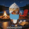 A tall iced beverage with whipped cream sits outdoors on a table near a campfire and lake at night.