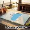 A rectangular glass cutting board printed with a Lake Tahoe graphic rests on a wooden bar overlooking a lake.