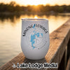 A white insulated wine tumbler featuring a blue map of Minong Flowage, Wisconsin, and a compass rose design, sitting on a wooden dock at sunset.