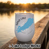 A white insulated tumbler with a custom Sand Lake Ontario map design sits on a wooden dock overlooking a lake at sunset.