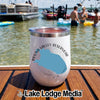 A white insulated travel tumbler with a compass and Round Valley Reservoir graphic sitting on a wooden boat deck with people swimming in the lake behind it.