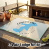 A glass cutting board featuring a Canobie Lake New Hampshire map rests on a wooden dock with drinks.