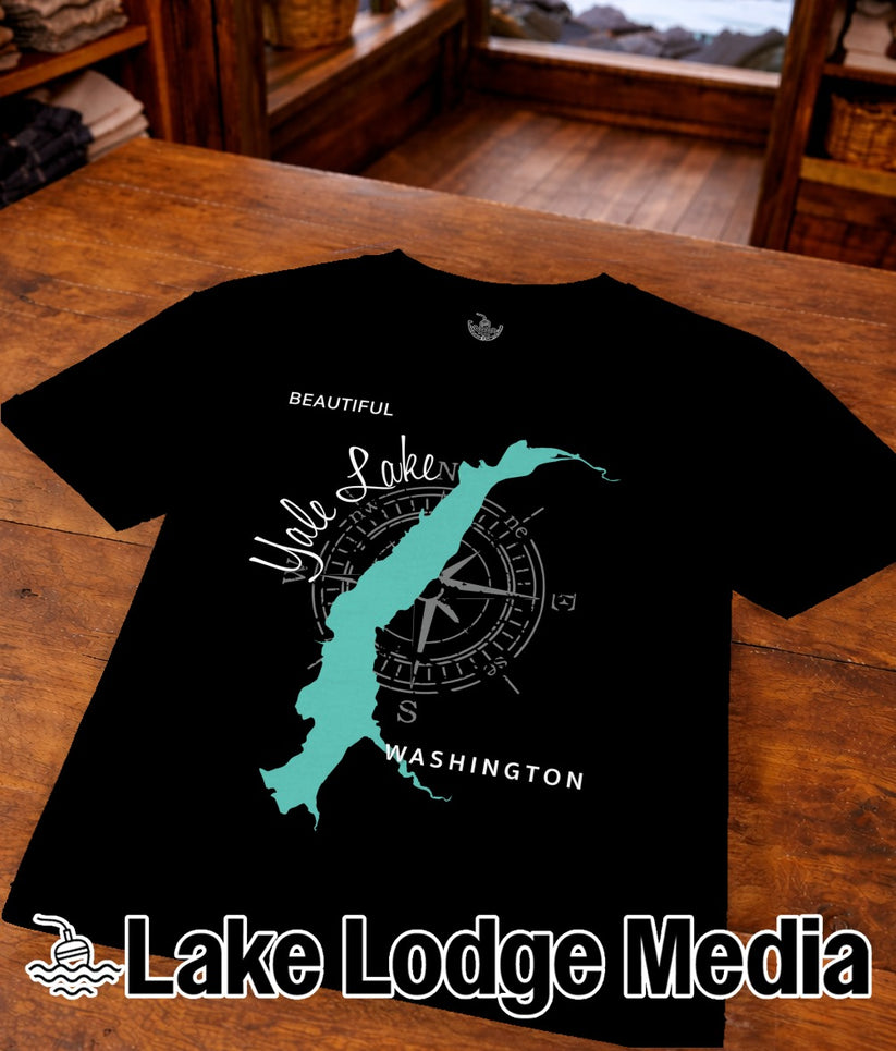 Black t-shirt with a turquoise lake outline and compass on a wooden table.