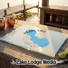 A frosted glass cutting board with a blue Pearl Lake, Wisconsin map design rests on a wooden dock beside copper mugs and lime slices.