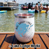 A white insulated tumbler featuring a Big Sandy Lake map graphic resting on a wooden boat deck with people swimming in the background.