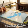 A glass cutting board featuring a blue map of Eagle Lake, California rests on an outdoor wooden table with copper mugs and limes nearby.