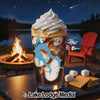 Glass of iced coffee with whipped cream, campfire, and lake scene in background.