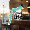 Turquoise map outline sticker with "Baker Life" text, on a glass surface.