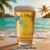 Beer glass with "Ottely Lake" design and compass, on a beach with ocean background.