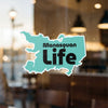 Teal Manasquan Life sticker on a glass surface with blurred interior background.