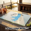 A glass cutting board featuring a blue map of Flathead Lake, Montana, sits on an outdoor wooden table near copper mugs and a dock.