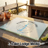 A rectangular glass cutting board rests on a wooden dock with Flaming Gorge Lake Wyoming printed over a compass design.