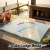 A rectangular glass cutting board with Lake George map design sits on a wooden dock beside copper mugs.
