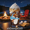 Glass of iced beverage with whipped cream, campfire, lake, and s'mores ingredients.