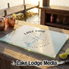 A glass cutting board featuring a compass design and Lake Oahe, South Dakota, rests on a wooden dock with drinks.