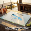 A rectangular glass cutting board rests on a wooden bar overlooking a sunny lake.