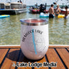 White stainless steel tumbler with a compass graphic and Hemlock Lake New York text, sitting on a wooden boat deck with people in the water in the background.