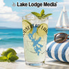 Tall glass with drink, lime, and mint on a beach with sailboat and sunglasses.