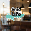 "Enid Life" text on a teal map of Oklahoma sticker, seen through glass.