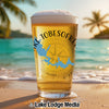 A pint glass filled with beer stands on a sunny beach, featuring a blue map of Lake Tobesofkee and a compass design.