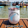 A white insulated wine tumbler sits on a wooden boat deck with a lake and people in the background.