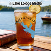 Tall pint glass filled with iced tea and a lemon slice, printed with a blue lake map graphic, sitting on an outdoor wooden table with a lake and boat in the background.