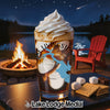 A glass with whipped cream and coffee sits by a campfire with s'mores ingredients.